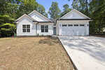 A modern single-story white house with black trim, attached two-car garage, concrete driveway, and minimal landscaping, surrounded by tall green trees under a clear blue sky.