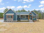 A modern single-story house with blue siding, brick accents, and wooden shutters. The home has a front porch with steps, surrounded by minimal landscaping, and sits on a large, bare lot with trees in the background.