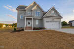 A newly built, two-story gray house with white trim, featuring a front porch, steps, attached two-car garage, and a driveway. The yard is covered with straw and small shrubs, under a partly cloudy sky.
