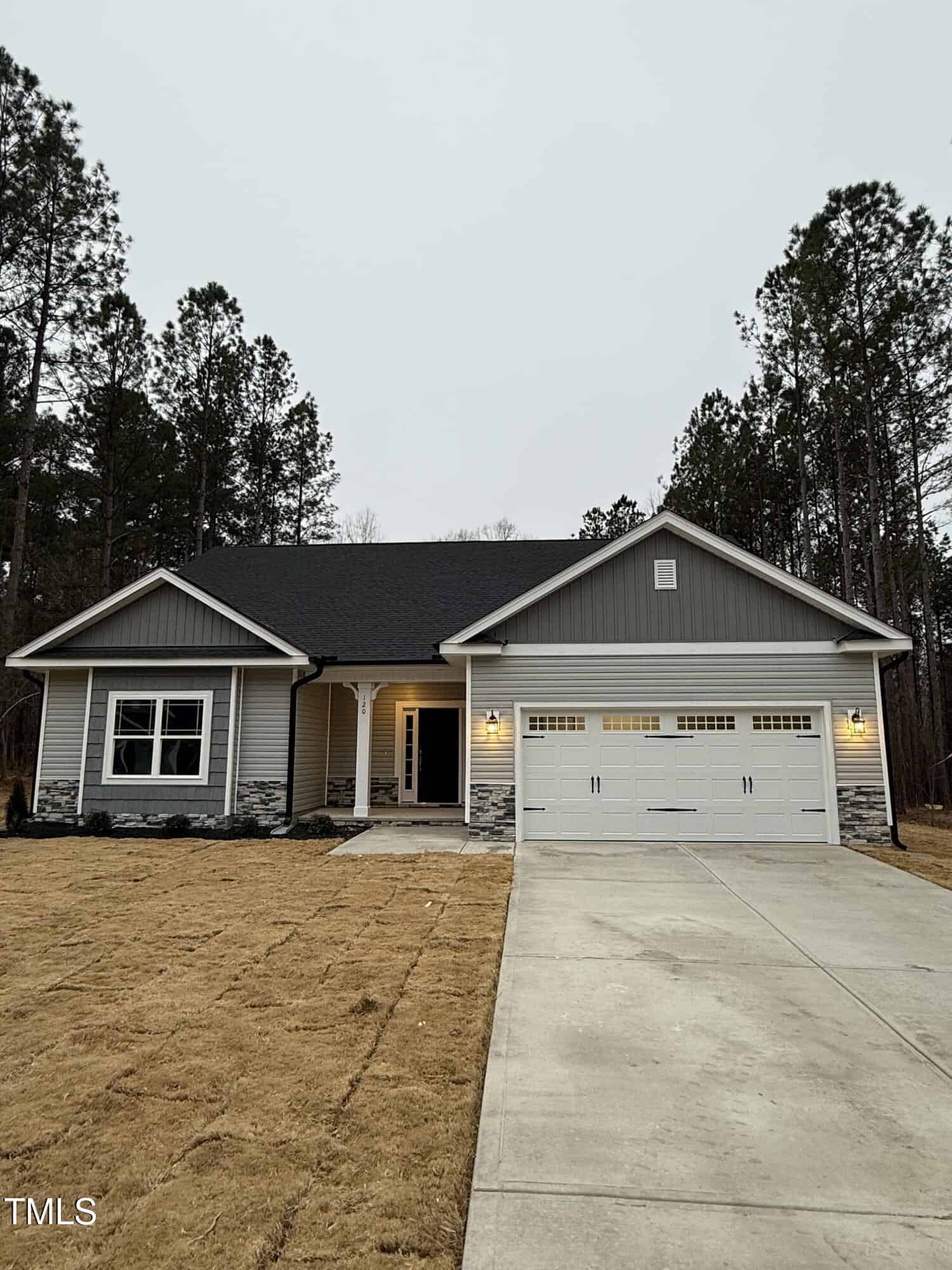 Single-story gray house with white trim, stone accents, and a two-car garage. The home has a covered front porch, lit entryway, and sits on a large lawn with trees in the background under an overcast sky.