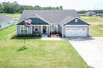 A modern single-story gray house with white trim, a front porch, and attached two-car garage. Two flags are displayed in the front yard, and the house is surrounded by a green lawn and neighboring homes.