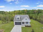 A modern, two-story house with a gray roof and stone accents sits on a large, grassy lot bordered by trees and woods under a blue sky with scattered clouds. A double driveway leads to an attached garage.