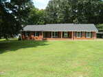 A single-story brick house with a dark roof, white trim, multiple windows, and a covered front porch, surrounded by a large, neatly mowed lawn and mature trees.