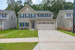 Two-story beige house with blue shutters, double garage, and a wide concrete driveway. Well-kept green lawn, minimal landscaping, and neighboring similar homes visible on both sides. Trees in the background.