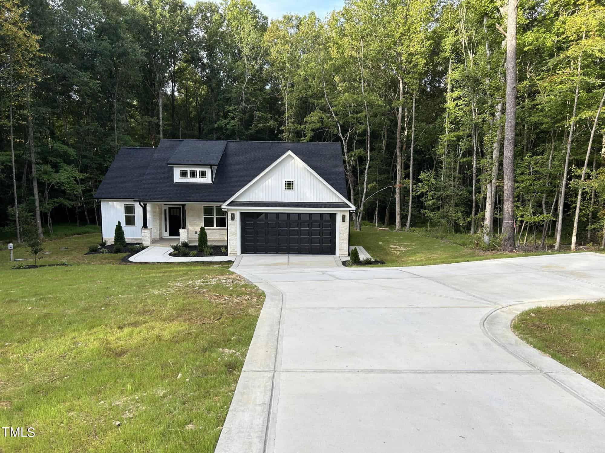 A modern white house with black trim and a black garage door sits at the end of a curved concrete driveway, surrounded by a grassy yard and tall, dense trees.