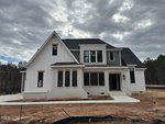 A newly constructed, modern farmhouse-style home with white siding, dark roof shingles, and large windows, sits on a bare, dirt lot with a concrete walkway, surrounded by trees under a cloudy sky.