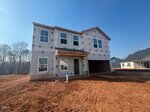 A two-story house under construction with exterior sheathing visible, window frames installed, and no exterior siding completed. The ground is bare, reddish dirt, and other homes are visible in the background under a clear sky.