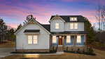 Two-story modern farmhouse with white siding, black roof, brick foundation, wooden front door, and a covered front porch. The house is surrounded by a paved driveway and minimal landscaping at sunset.