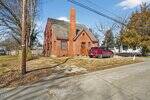 A red brick house with a steep roof and central chimney sits on a corner lot. A red SUV is parked on a gravel driveway next to the house. Leafless trees and utility poles are visible nearby.