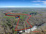 Aerial view of a forested area in early spring with a large, irregularly shaped plot of land outlined in red. A house and driveway are visible within the outlined boundary. Surrounding trees have sparse foliage.