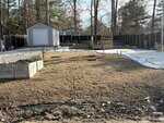 A backyard with brown grass, a raised garden bed on the left, a storage shed in the back left corner, a wooden deck in the center, and a black privacy fence surrounding the area. Trees are visible in the background.