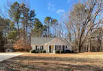 A single-story white house with a gray roof sits in front of a wooded area. There are large windows, bushes by the entrance, a concrete driveway, and leaf-covered ground under a blue sky.