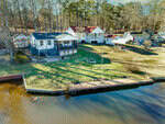 Aerial view of lakeside homes with docks, grassy yards, and trees in the background. The shoreline features a wooden retaining wall and a small pier extending over the water.