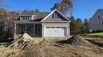 A newly constructed two-story house with gray siding and a white garage door sits behind two large piles of dirt in a yard with bare soil. Trees with fall foliage are visible in the background.