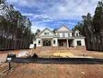 A two-story house under construction with a mix of brick and white siding exterior, surrounded by dirt, construction materials, and a black silt fence, with pine trees in the background.