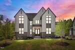 Two-story modern farmhouse with gray siding, black trim, and large windows, set against a colorful sunset sky. The house has a covered front entrance, landscaped yard, and trees on both sides.