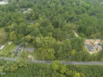 Aerial view of a suburban area with houses surrounded by dense green trees. A two-lane road runs horizontally across the bottom of the image, with homes, driveways, and open yards visible nearby.