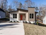 Single-story house with beige siding, white brick foundation, and an orange front door. There are three front windows, a small porch with railings, and a concrete driveway. The yard has some grass and shrubs.