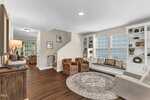 A living room with hardwood floors, built-in shelves and window seat, three armchairs around a circular rug, and a view of a hallway with stairs leading up. Large windows line one wall, letting in natural light.