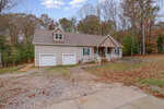 A tan, single-story house with a double garage, front porch, and gabled roof sits among trees with fallen autumn leaves covering the yard and driveway.