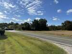 A two-lane road runs beside a grassy area and open field with trees in the background under a mostly clear blue sky with scattered clouds.