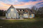 A modern two-story house with light blue siding, a dark gray roof, and large windows, set on a grassy lawn with minimal landscaping, photographed at sunset with a colorful sky.