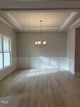 A dining room with light wood flooring, beige walls, white wainscoting, a coffered ceiling, and a five-light chandelier hanging from the ceiling. Sunlight streams in through large windows on the left.
