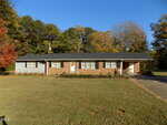 A single-story ranch-style house with a mix of brick and light siding, dark shutters, and a covered front porch. The house is surrounded by grass and trees with some autumn foliage. A driveway runs along the right side.