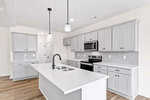 Modern kitchen with white walls, light gray cabinets, stainless steel appliances, a white island with a sink and black faucet, pendant lighting, and light wood flooring.