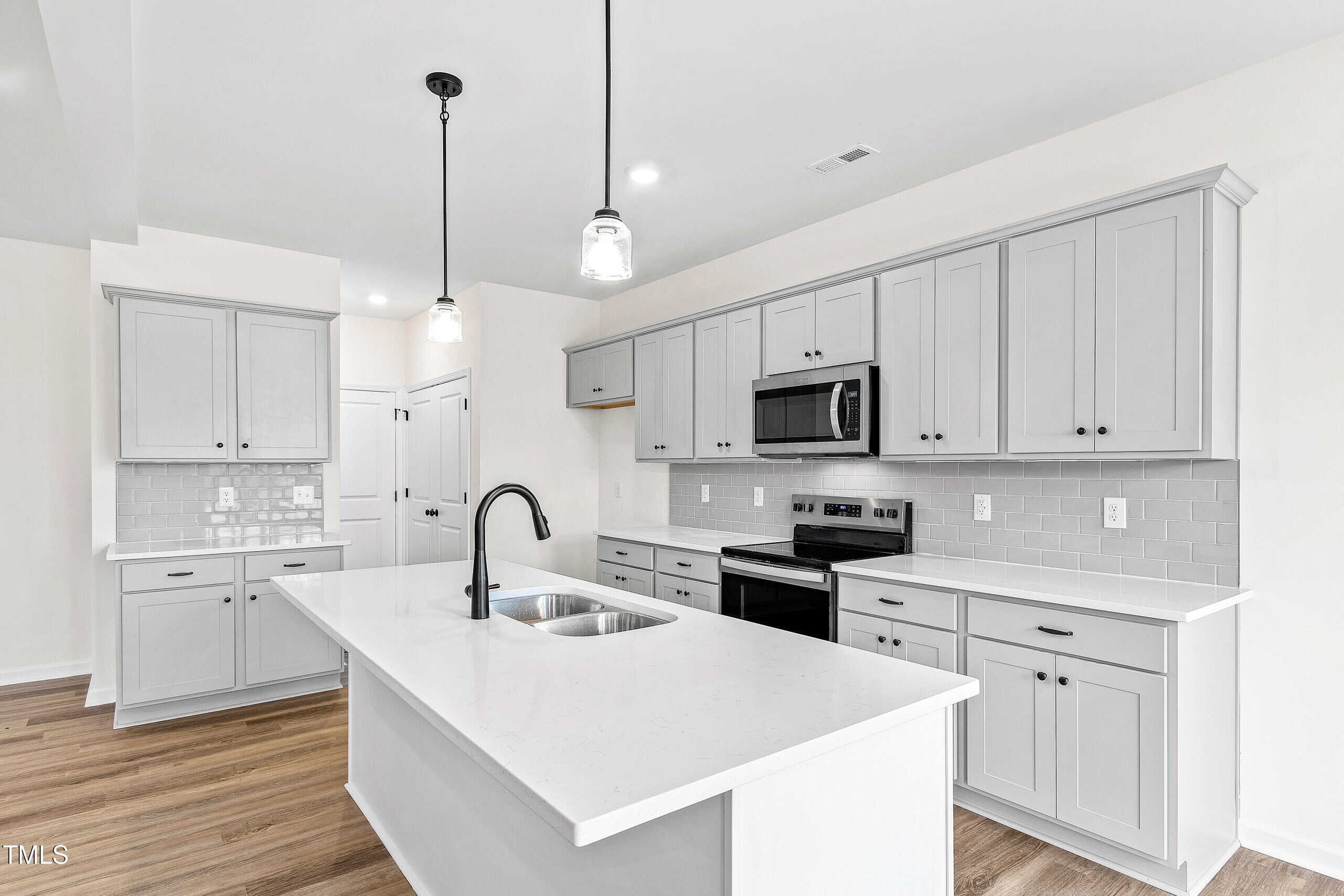 Modern kitchen with white walls, light gray cabinets, stainless steel appliances, a white island with a sink and black faucet, pendant lighting, and light wood flooring.