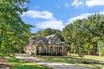 A two-story brick house with a front porch, American flag, and landscaped yard sits among trees. A driveway leads to a three-car garage. The sky is blue with scattered clouds.