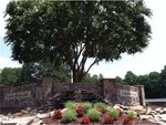 A stone sign for Lake Royale is set in front of a large tree, with landscaping that includes red flowers, rocks, and a small stone waterfall. The background shows lush green trees under a clear sky.
