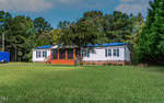 Single-story white house with a brick foundation, front porch with wooden railings, and solar panels on the roof, surrounded by grass and trees under a partly cloudy sky.