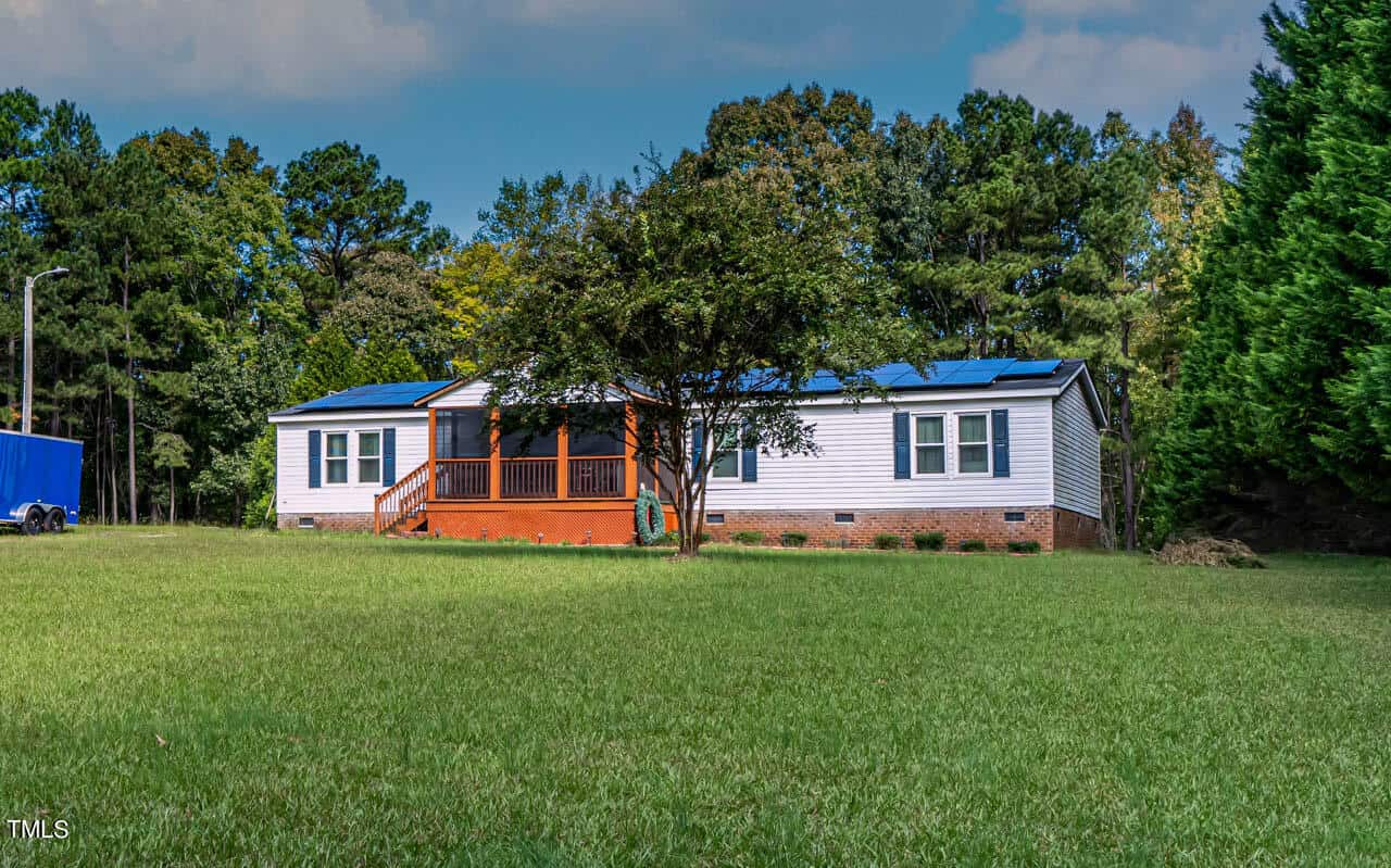 Single-story white house with a brick foundation, front porch with wooden railings, and solar panels on the roof, surrounded by grass and trees under a partly cloudy sky.