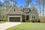 A two-story gray house with white trim, a dark gray double garage, and a small front porch with black railing. The house has a large driveway and a well-kept lawn, surrounded by tall trees and landscaping.