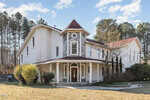 A large, two-story house with white exterior walls, a prominent turret, red roof, and wraparound porch. The house is surrounded by trees and bushes, with a mostly bare lawn in front.
