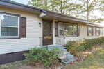 A single-story beige house with brown shutters and trim, featuring a front entrance with steps, a glass door, metal railings, and several large windows. Bushes line the front, and tall pine trees are visible in the background.