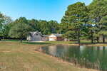 A house and detached garage sit among trees near a small pond, with a grassy yard in the foreground and a clear blue sky overhead.
