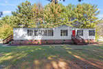 A single-story manufactured home with gray siding, a red front door, multiple windows, and a brick foundation, surrounded by green grass, trees, and a clear blue sky.