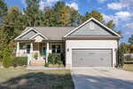 Single-story beige house with a covered front porch, green accents, double garage, and neatly trimmed shrubs. A concrete driveway leads to the garage. Trees with autumn foliage are visible in the background.