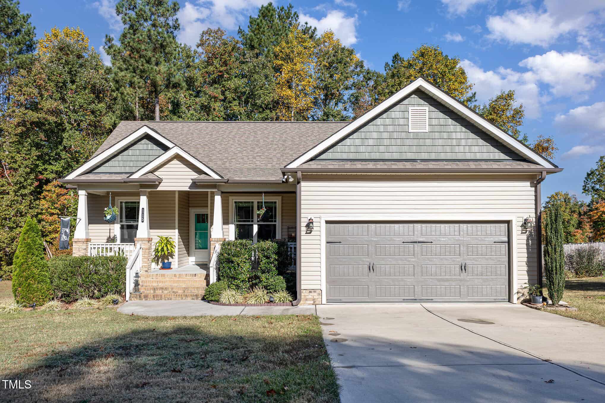 Single-story beige house with a covered front porch, green accents, double garage, and neatly trimmed shrubs. A concrete driveway leads to the garage. Trees with autumn foliage are visible in the background.