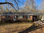 A single-story brick house with a dark shingle roof, front door, several windows, and a small front porch. There are shrubs along the front and a carport on the right side. Leafless trees cast shadows on the lawn.