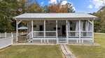 A white single-story house with a metal roof and a covered front porch sits on a grassy lawn. Trees with autumn foliage are in the background. Steps lead up to the porch, which has railings and outdoor lights.