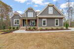 A modern two-story house with gray siding, white trim, and a covered front porch. The yard features new landscaping with small shrubs and mulch, and the driveway curves in front of the home.