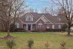 A two-story brick house with white trim, multiple gables, and a covered front porch. The front yard has green grass, small shrubs, and several large leafless trees.