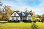 A two-story house with white siding and black trim sits on a large, well-maintained lawn. The yard has a few young trees, and the house is surrounded by trees with autumn foliage under a blue sky.