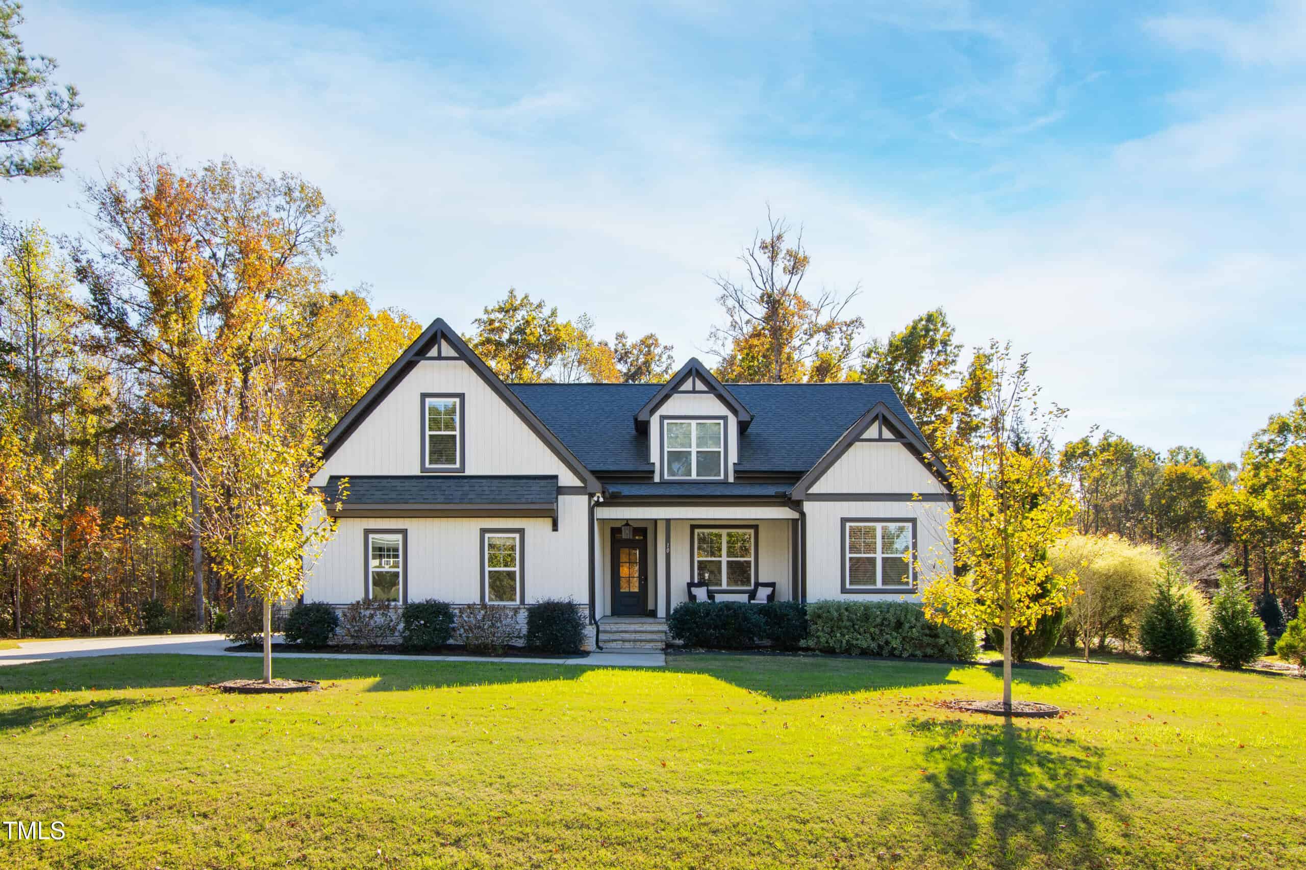 A two-story house with white siding and black trim sits on a large, well-maintained lawn. The yard has a few young trees, and the house is surrounded by trees with autumn foliage under a blue sky.