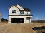 A two-story house under construction with house wrap covering the exterior, open garage and window frames visible, surrounded by dirt and gravel, against a clear blue sky.