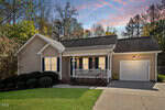 Single-story beige house with black shutters, a covered front porch, attached one-car garage, manicured bushes, and concrete driveway. Trees surround the property, and the sky shows a colorful sunset.