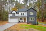 Two-story modern house with white and dark gray siding, a double garage, and a front porch with two chairs. The house is surrounded by trees and has a grass lawn with a concrete driveway.